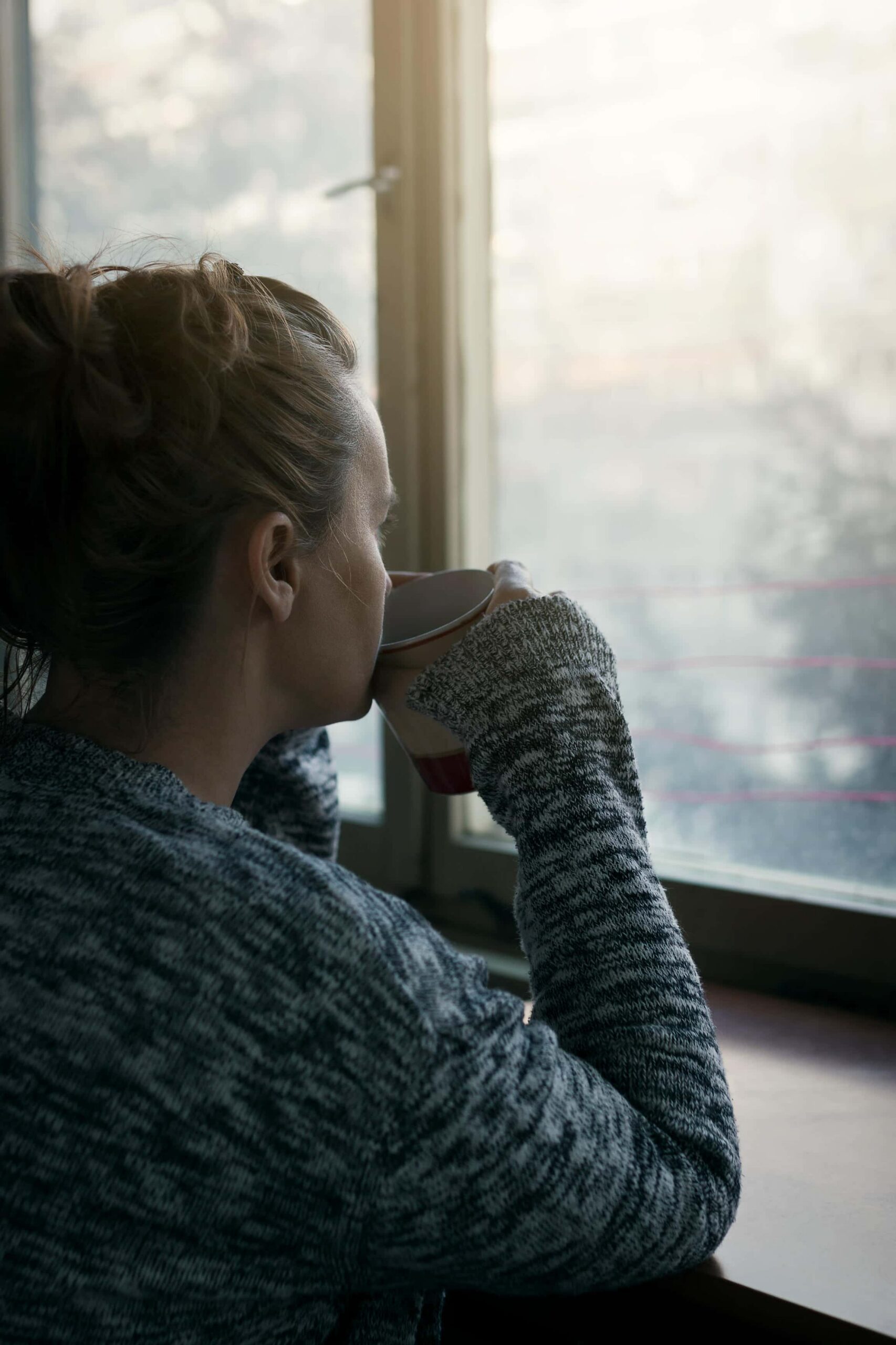 Sad woman looking out a window while holding a cup of coffee and exploring alternatives to alcohol for anxiety with support from Flatirons Recovery.